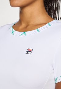 Detailný záber osoby v bielom športovom tričku s vzorovaným výstrihom a logom Fila na hrudi.