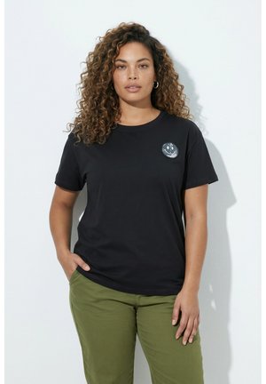 Kroeshaarige vrouw die een zwart T-shirt draagt met een smiley-patch en groene broek, staande met één hand in de zak tegen een witte muur.