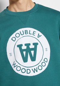 Tyrkysová bavlněná mikina s kulatým bílým logem obsahujícím text "DOUBLE A WOOD WOOD" a stylizované iniciály "W" uprostřed.