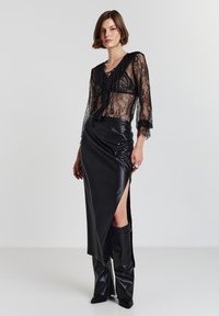Blusa nera in pizzo con maniche a campana, stratificata su una gonna in pelle nera con spacco alto. Indossata con stivali in pelle liscia al ginocchio.
