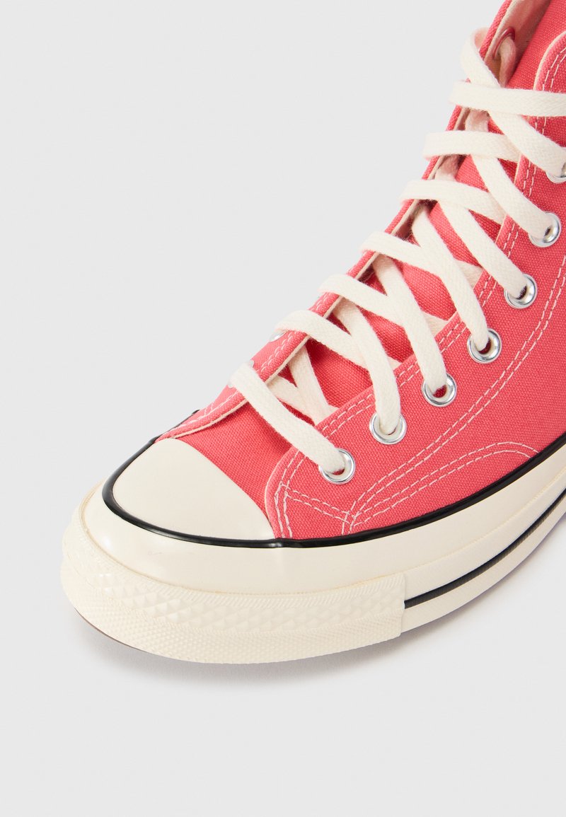 Rød højt klippet sneaker i lærred med hvide snørebånd, en gummiforfod, sort kant og metaløjer. Tekstureret side med syede detaljer.