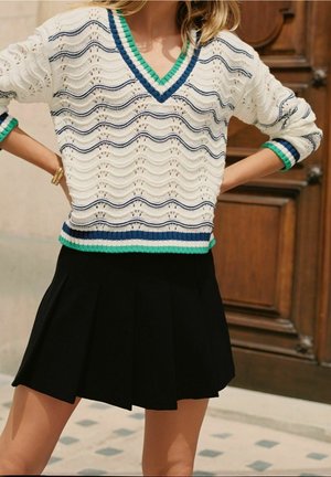 Personne portant un pull en tricot blanc avec des rayures bleues et vertes et une jupe plissée noire, debout avec les mains sur les hanches près d'une porte en bois.