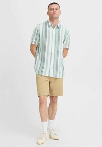 Chemise à manches courtes, boutonnée, de couleur vert clair avec des rayures verticales, associée à un short beige et des chaussettes blanches, offrant une coupe décontractée et un tissu léger.