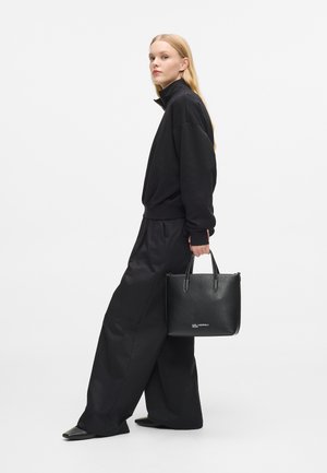 Donna con i lunghi capelli biondi, vestita con abiti neri larghi, che tiene una borsa a tracolla in pelle nera con del testo bianco piccolo, mentre cammina di lato.