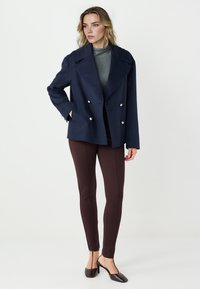 Cappotto blu navy doppiopetto con bottoni dorati, abbinato a leggings marroni e scarpe slingback con tacco marrone, indossato sopra una maglia grigia.