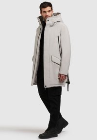 Parka lung de culoare gri deschis, cu glugă, închidere cu fermoar frontal, două buzunare laterale și accente negre. Asortat cu pantaloni cargo negri și cizme.