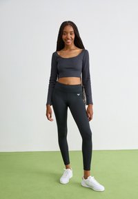 Dark gray cropped long-sleeve top with ribbed texture, paired with black leggings and white sneakers. Figure-hugging fit with logo detail.