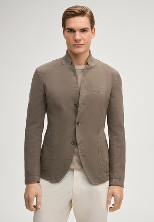 Junger Mann stehend, trägt ein taupefarbenes, geknöpftes Sakko über einem beigen Hemd mit weißen Hosen, neutraler Hintergrund.