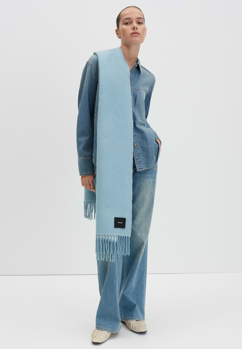 Sciarpa azzurra con frange, realizzata in un materiale soffice, indossata sopra una camicia di jeans e jeans a gamba larga, con un piccolo logo nero.