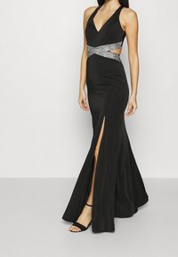 Černé večerní šaty s halter krkem, hlubokým výstřihem do V, vykrojenými boky, opaskem s rhinestony a vý poutavým rozparkem až k stehnům. Hladká, přiléhavá látka.