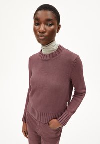 Strickpullover in gedämpftem Mauve mit geripptem Kragen und Ärmeln, ausgestattet mit einem strukturierten Oberteil und schlankem Schnitt, kombiniert mit einem T-Shirt mit hohem Kragen darunter.