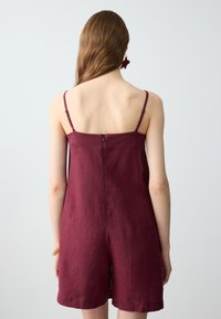 Bordeaux linnen jumpsuit, mouwloos met dunne bandjes, voorzien van een ritssluiting aan de achterkant en een relaxte pasvorm, eindigend halverwege de bovenbenen.