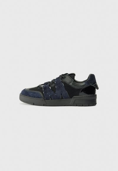 Zwarte en marineblauwe sneakers zijn gemaakt van suede en mesh materialen, met een dikke zool en een opvallend uitgesneden "M" detail aan de zijkant.