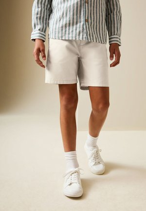 Person trägt beige Shorts, weiße Socken, weiße Turnschuhe und ein blau-weiß gestreiftes Langarmshirt und steht auf einem hellen Boden.