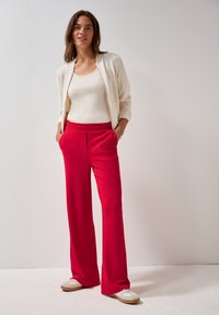 Červené široké kalhoty s texturovaným designem mají boční kapsy, v kombinaci s krémovým topem na ramínka a lehkou bomber bundou. Bílé boty dokončují outfit.