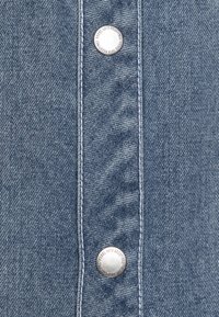 Denimstoff in Hellblau, mit einer vertikalen Knopfleiste mit vier metallischen Druckknöpfen und kontrastierenden weißen Nähten.