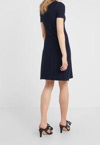 Abito navy a maniche corte con tessuto testurizzato e gonna svasata. Presenta un motivo di cucitura sottile. Il modello indossa sandali neri con tacco e un dettaglio a fiocco.