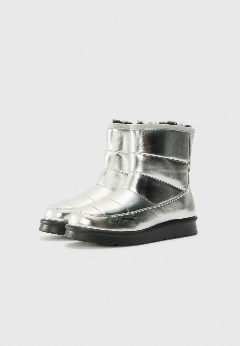 Bottines métalliques argentées avec une texture lisse, un design rembourré et une semelle en caoutchouc noire. Présente le logo de la marque sur le côté.