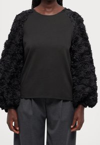 Haut noir avec des manches en dentelle florale texturée, col rond et coupe décontractée. Corps en tissu lisse, volume dans les manches, design minimaliste.