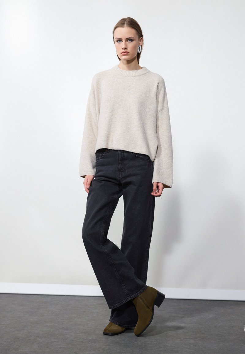 Suéter oversized color beige claro con un cuello acanalado, combinado con unos jeans oscuros de cintura alta. Botas de tobillo verde oliva completan el look.