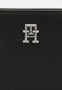 Black leather wallet featuring a textured surface and a silver metallic logo. Design includes a zip closure and minimalist accents.