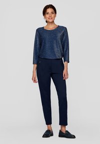 Marineblauer, glitzernder Langarmoberteil, kombiniert mit schmal geschnittenen marineblauen Hosen. Glattes Material mit Fronttaschen; getragen mit dunklen Slippern.