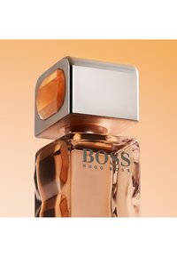 Przezroczysta szklana butelka perfum z bursztynowym płynem, dużą srebrną nakrętką z bursztynowym kamieniem, oznaczona napisem "BOSS HUGO BOSS" na ciepłym beżowym tle.