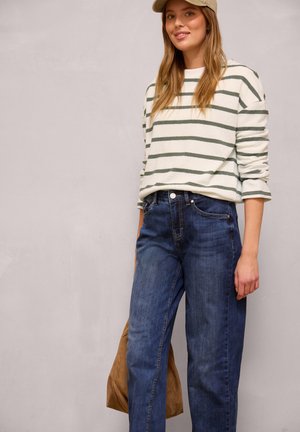 Junge Frau mit beiger Mütze, weiß-grün gestreiftem Langarmshirt, blauer Jeans und einer braunen Tasche vor neutralem Hintergrund.