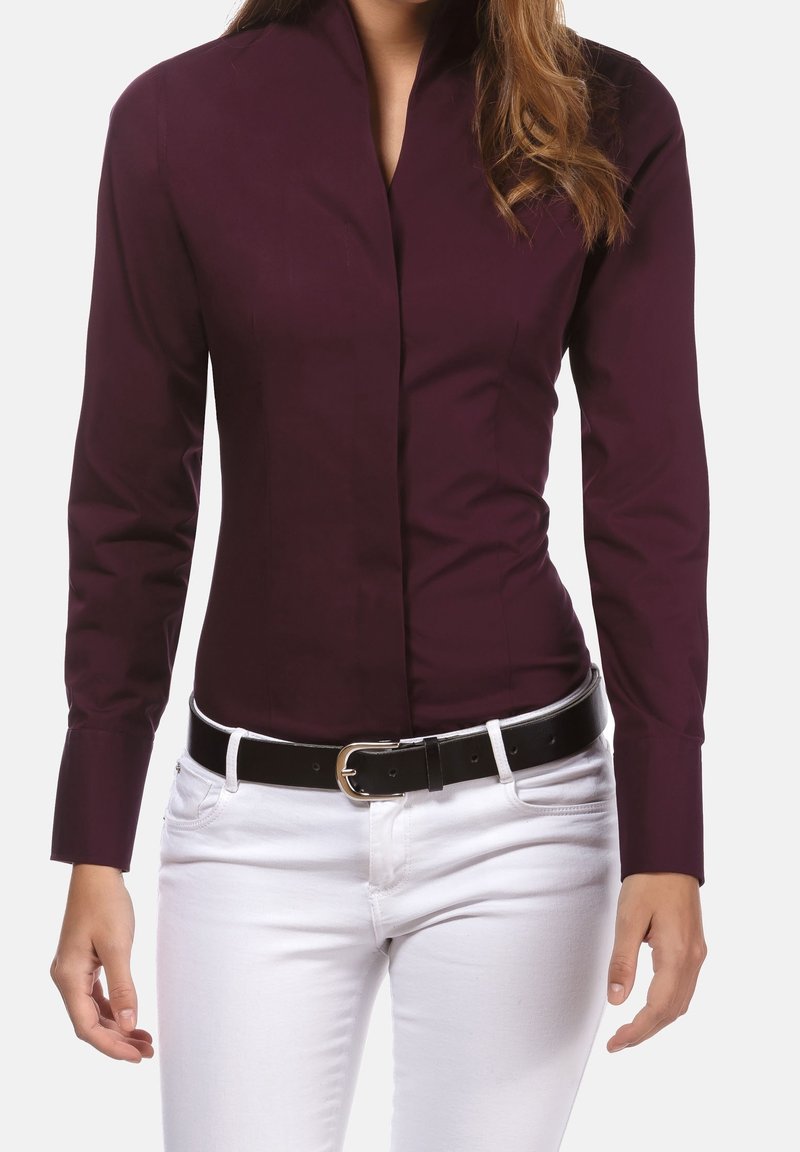 Femme portant une chemise ajustée bordeaux à manches longues rentrée dans un pantalon blanc avec une ceinture noire, debout devant un fond uni.