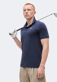 Joven con una camisa polo azul marino y pantalones beige, sosteniendo un palo de golf sobre el hombro con un guante blanco en una mano.