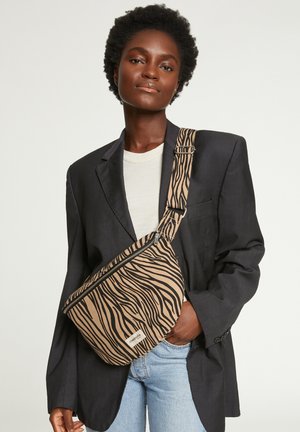 Sac en bandoulière beige avec des rayures zèbre noires, doté d'une fermeture éclair et d'une bandoulière ajustable. Porté par-dessus un t-shirt clair et un blazer.