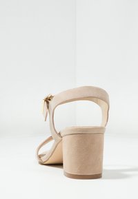 Beige mokkasini nahkasaappaat, joissa on nilkkaremmi ja kultainen solki. Tasainen pinta, avoin takapuoli ja pyöristetty kärki.