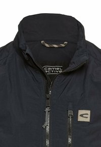 Granatowa, lekka kurtka z wysokim kołnierzem, zamkiem błyskawicznym z przodu i teksturowanym materiałem. Posiada małą naszywkę z logo oraz wzorzystą sznurówkę.