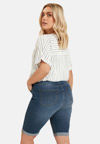Denim bermuda shorts with blue finish, rolled hem, and back pockets, paired with a white striped blouse featuring a relaxed fit.