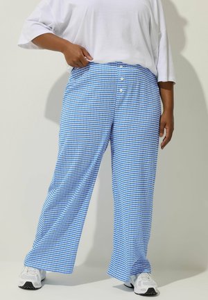 Persona vistiendo pantalones sueltos a cuadros azules y blancos con botones, una camisa blanca de gran tamaño y zapatillas blancas, de pie contra un fondo liso.