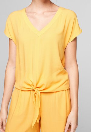 Nainen, joka pukeutunut lyhythihaiseen, V-kaulaiseen keltaiseen toppiin, joka on solmittu vyötärölle, ja hänellä on päällään yhteensopivat keltaiset housut yksinkertaista taustaa vasten.