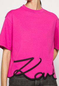 Fuchsia Baumwoll-T-Shirt mit Rundhalsausschnitt, kurzen Ärmeln und einem schwarzen gestickten "Love"-Design entlang des Saums. Weiche Textur.