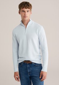 Svetlomodri pulover z zadrgo do četrtine dolžine, z reliefnim pletenjem, rebrastimi manšetami in spodnjim robom. Nošen čez modre kavbojke, z majhnim logotipom na pasu.