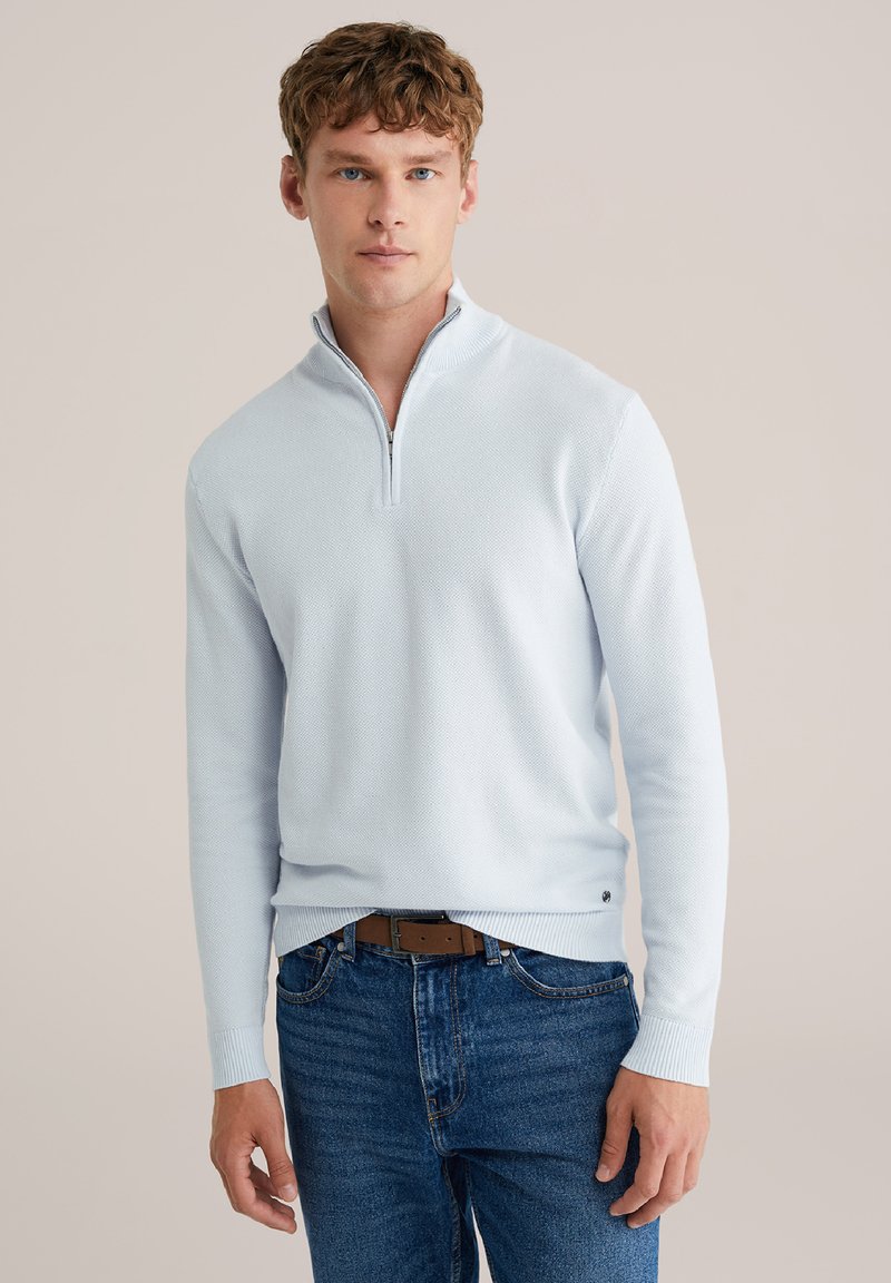 Svetlomodri pulover z zadrgo do četrtine dolžine, z reliefnim pletenjem, rebrastimi manšetami in spodnjim robom. Nošen čez modre kavbojke, z majhnim logotipom na pasu.