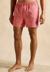 Muž oblečený v korálovo ružových plavkách s viazaním v páse a malým námorníckym modrým logom na ľavom stehne, stojí bosý na vzorovanej podlahe.