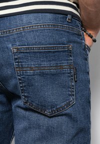 Un jean en denim bleu foncé présente un design de poche arrière avec des coutures contrastantes et une étiquette de marque tissée. Le tissu a une texture classique.