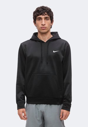 Jeune homme aux cheveux bouclés et foncés portant un sweat à capuche noir Nike et un pantalon gris clair, debout devant un fond clair uni.