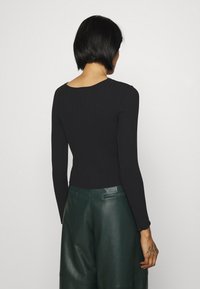 Schwarzes, geripptes Langarmshirt mit rundem Ausschnitt, kombiniert mit grünen, weiten Lederhosen. Der Stoff sieht glatt und figurbetont aus.