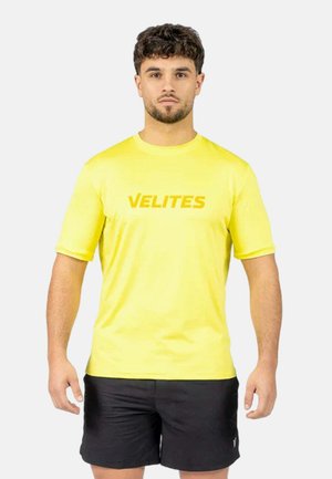 Hombre con pelo corto y rizado lleva una camiseta de manga corta de color amarillo brillante con el texto "VELITES" y pantalones cortos deportivos negros, de pie contra un fondo claro.
