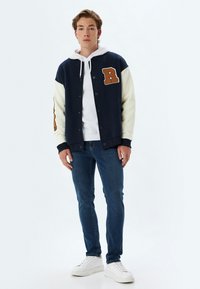 Chaqueta varsity en azul marino y crema con un parche texturizado en forma de letra 'R', combinada con una sudadera blanca, jeans azules y zapatillas blancas. Tejido suave, ajuste casual.