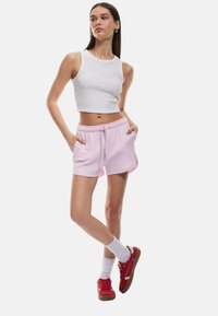 Top court côtelé blanc associé à un short rose texturé avec une taille élastique et des poches latérales. Le mannequin porte des baskets rouges et des chaussettes blanches.