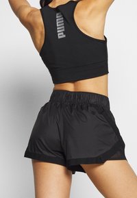 Mujer vistiendo un top deportivo negro de Puma y pantalones cortos atléticos negros con cintura elástica, posando con los brazos en alto frente a un fondo neutro.