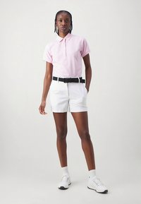 Ροζ ανοιχτόπουλο πόλο μπλουζάκι με γιακά και τέσσερα κουμπιά, συνδυασμένο με λευκά σορτς, μαύρη ζώνη και λευκά αθλητικά παπούτσια με λεπτομέρειες.