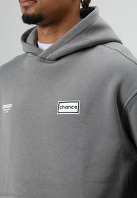 Grijze hoodie met een kangeroezak, met een wit "chance" logo binnen een rechthoekige omlijning en de tekst "STRONGER" aan de linkerkant.