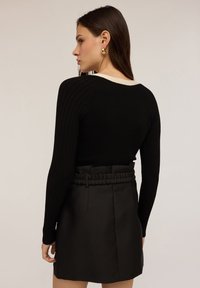 Jonge vrouw met lang donker haar draagt een zwarte geribbelde trui en een zwarte rok met hoge taille en doorlopende riem, terwijl ze zijwaarts staat tegen een effen achtergrond.